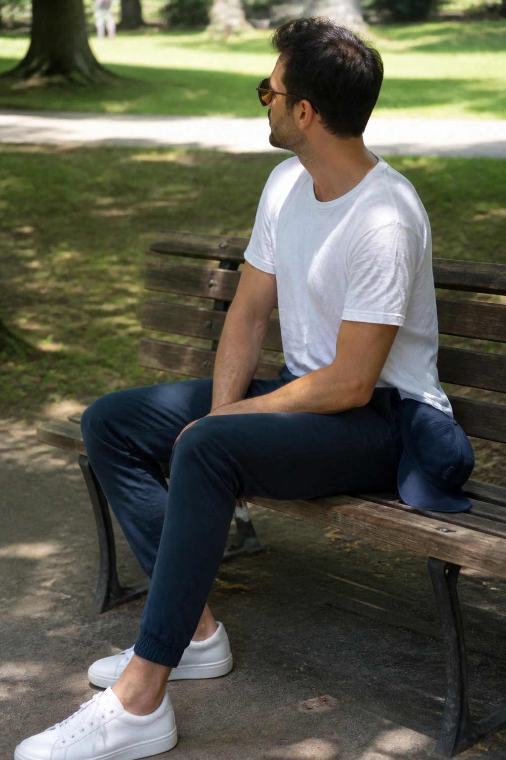 Man in white t-shirt and navy pants sits on park bench with blue cap nearby