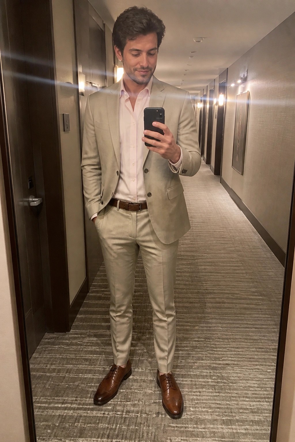 Man in beige suit and pink shirt taking mirror selfie in hotel hallway