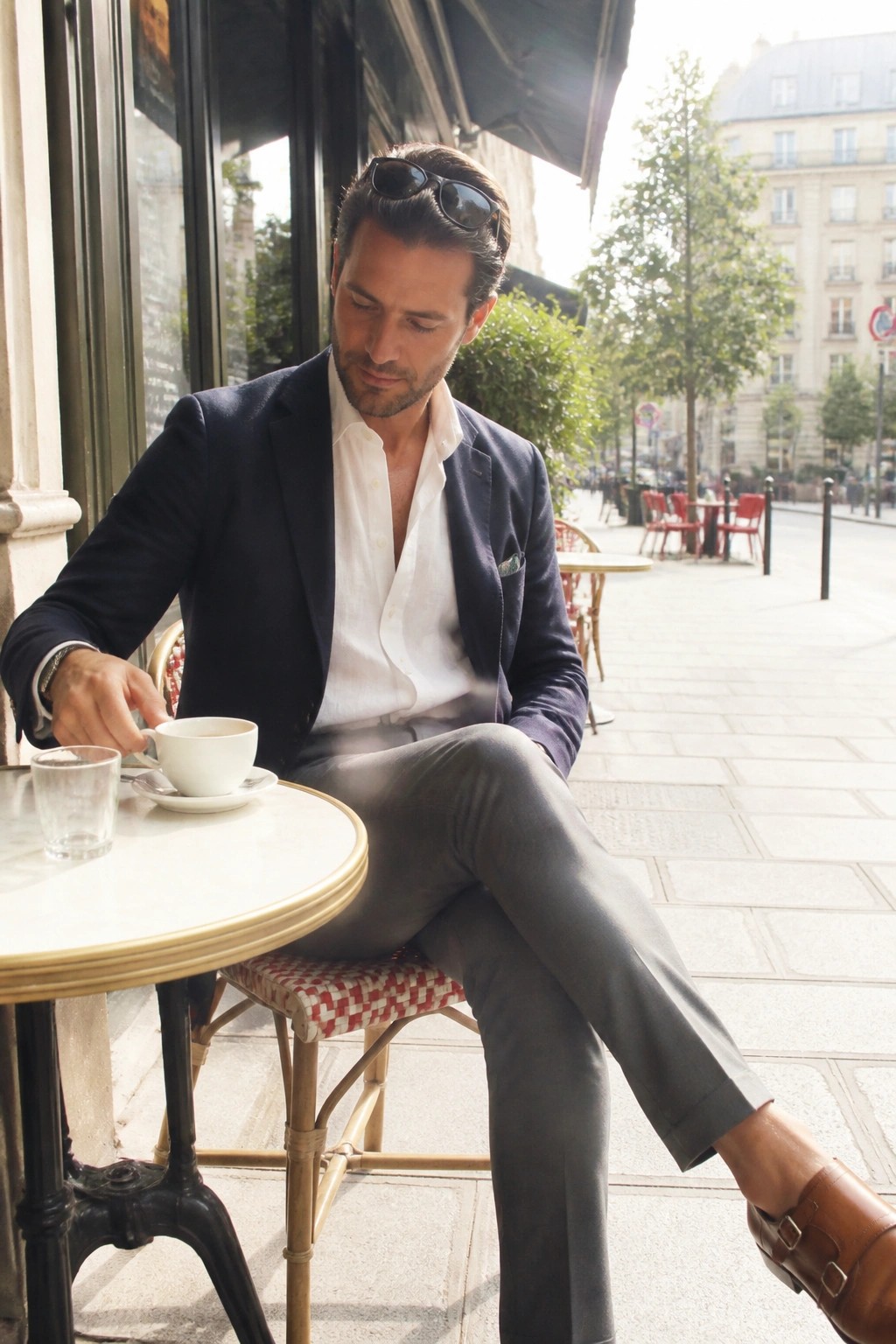 Man in navy blazer and white shirt sipping coffee at outdoor Parisian café.
