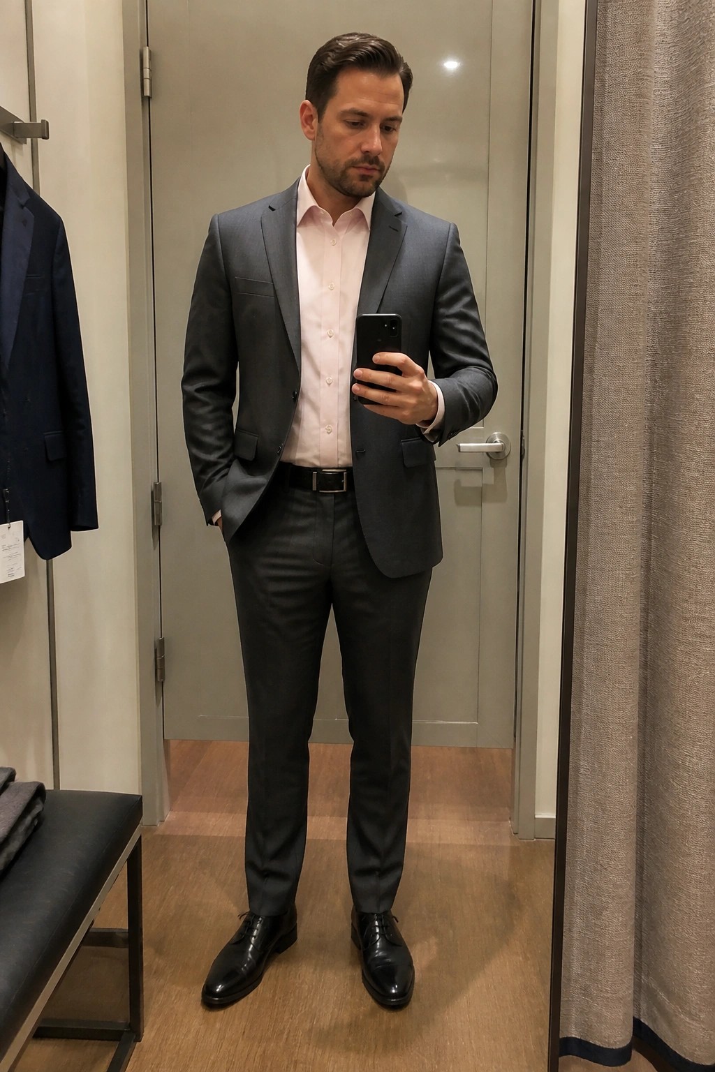 Man in gray suit and pink shirt taking mirror selfie in fitting room