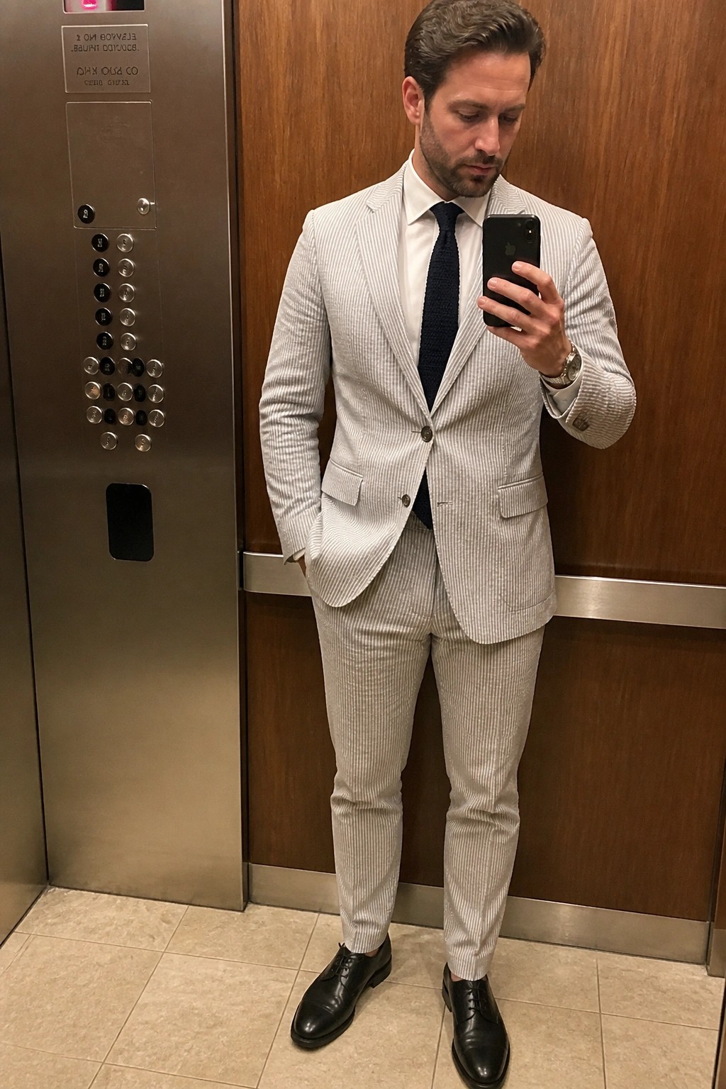 Man in light gray pinstripe suit taking mirror selfie in elevator