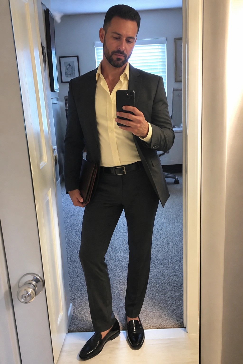 Man in gray suit and yellow shirt taking mirror selfie with phone and portfolio