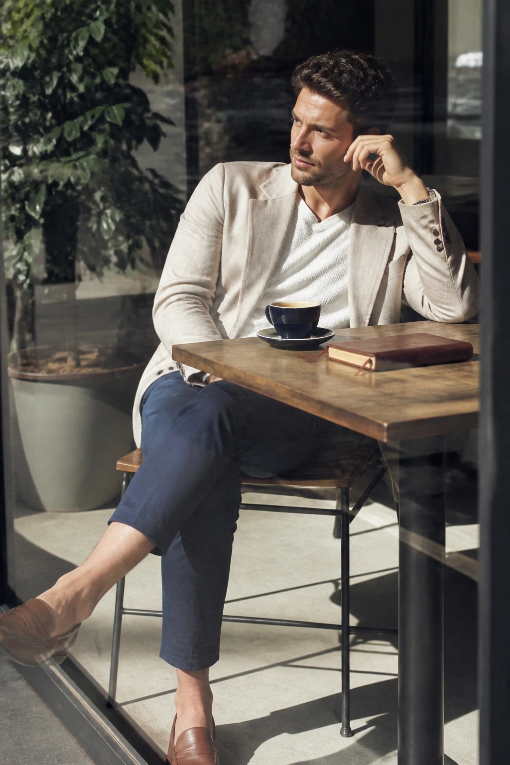 Man wearing beige blazer over white textured sweater, navy trousers, and brown loafers seated at wooden table with coffee cup and notebook.