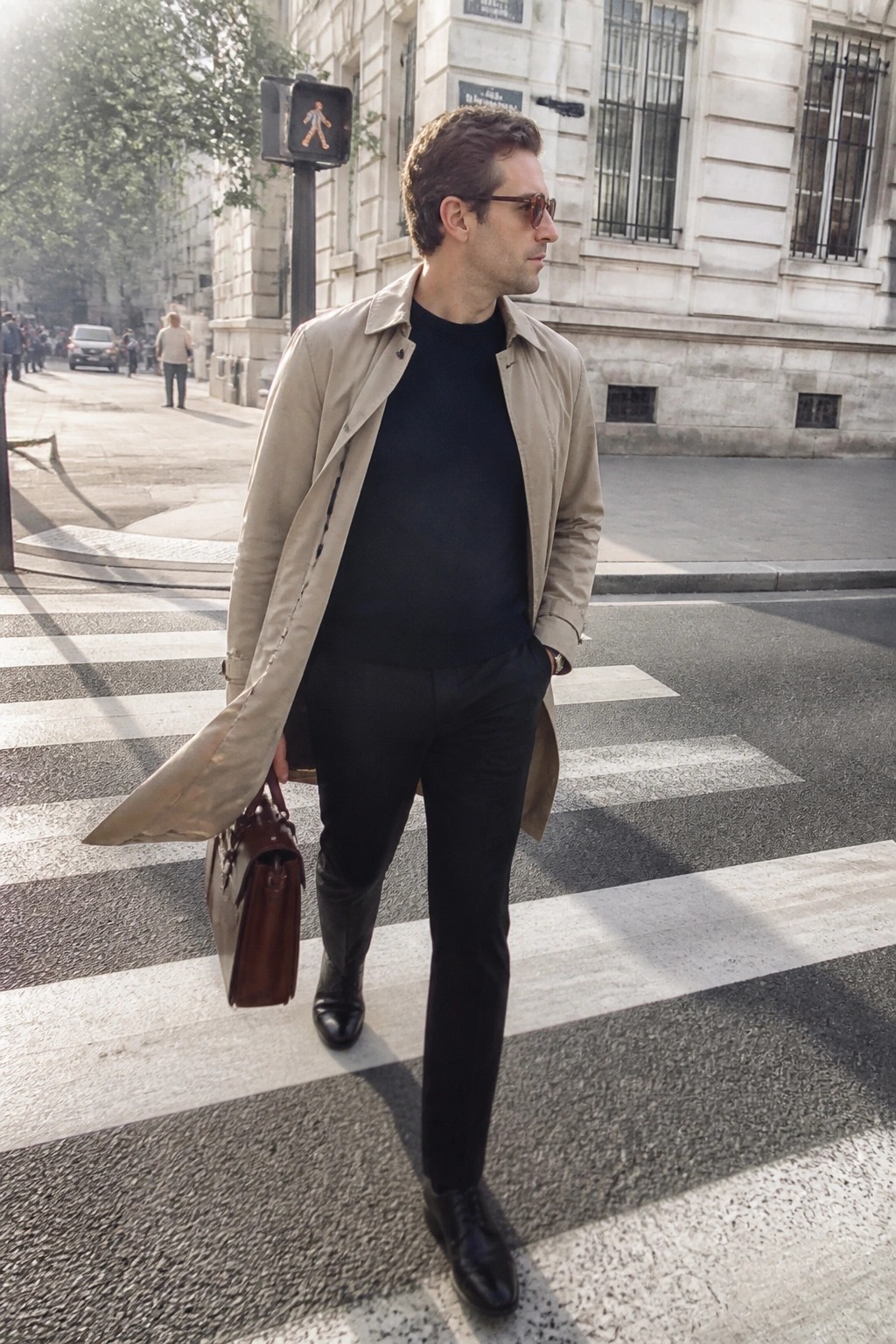 Man wearing open beige trench coat over black crewneck sweater and black trousers with black dress shoes while carrying a brown leather briefcase on a city crosswalk.