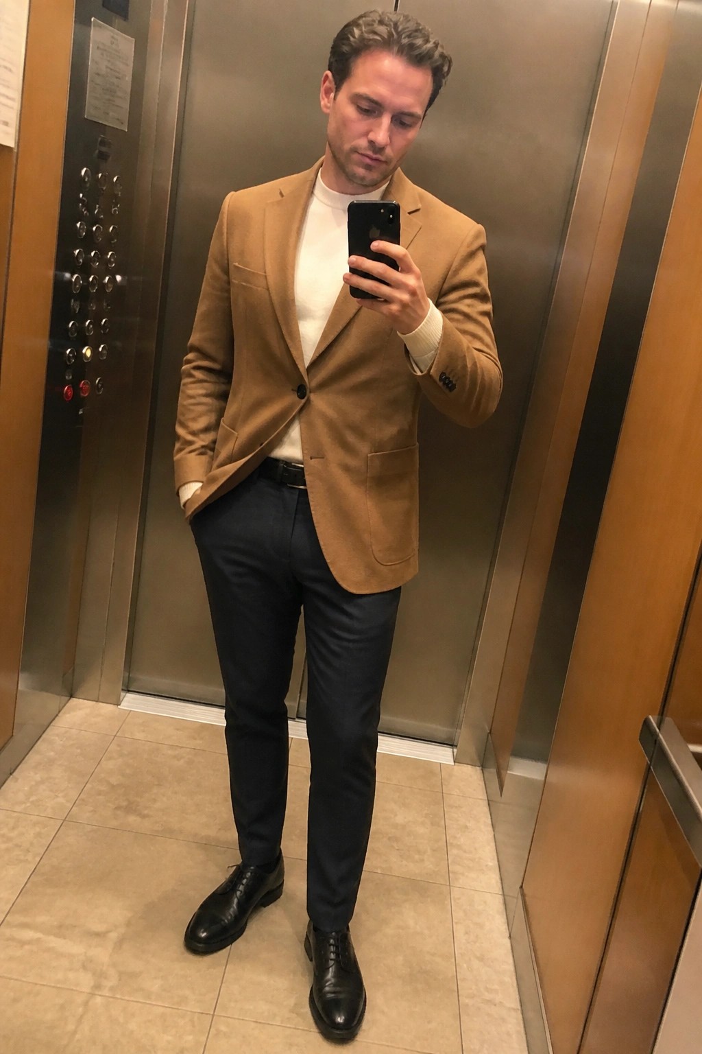 Man in tan blazer over white sweater with navy pants holding phone in elevator