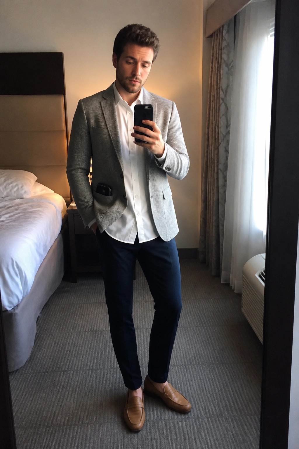 Man in gray blazer and white shirt takes mirror selfie in hotel room