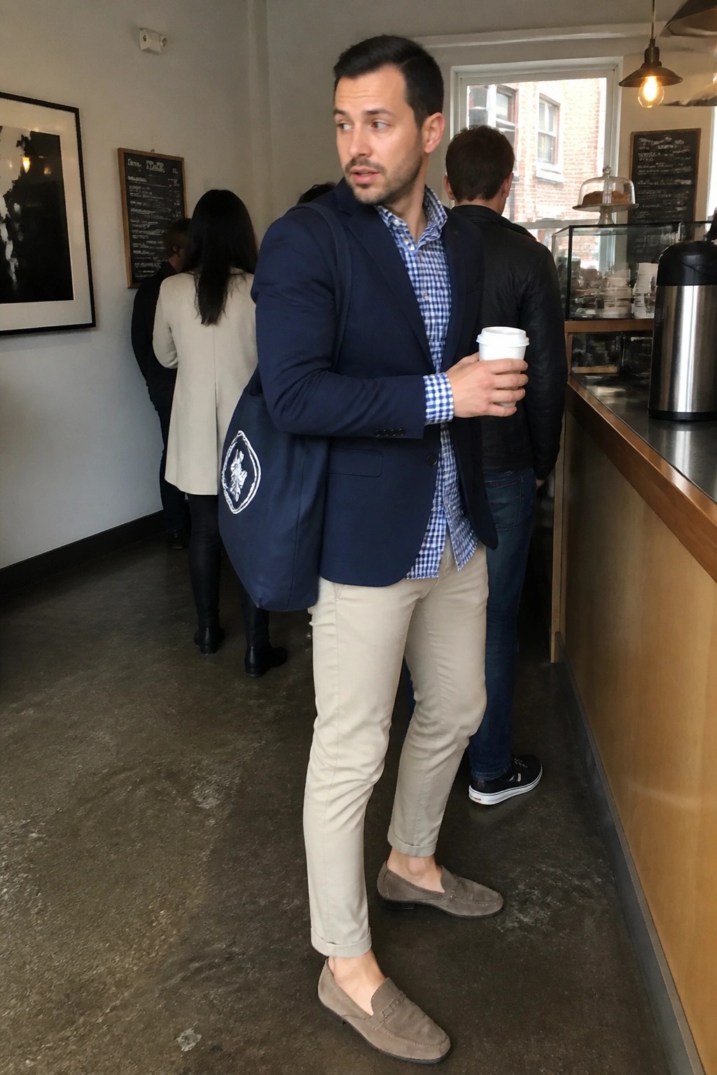 Man in navy blazer and checkered shirt holding coffee with tote bag in cafe