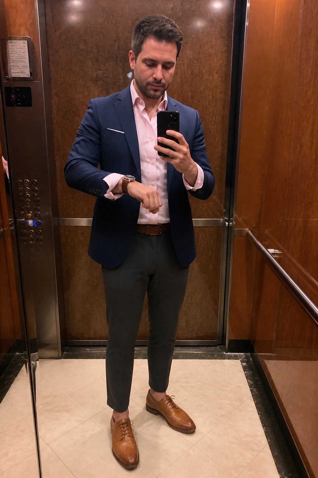 Man in navy blazer over pink shirt checks watch while holding phone in elevator