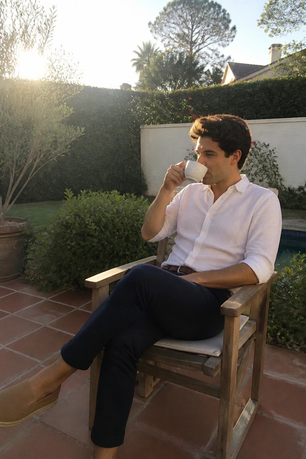 Man in white shirt and navy pants sipping from cup in wooden patio chair