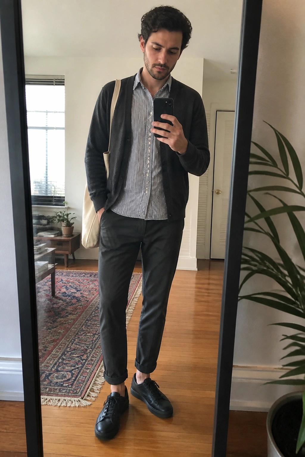 Man in striped shirt and dark cardigan holding phone with beige tote bag in mirror