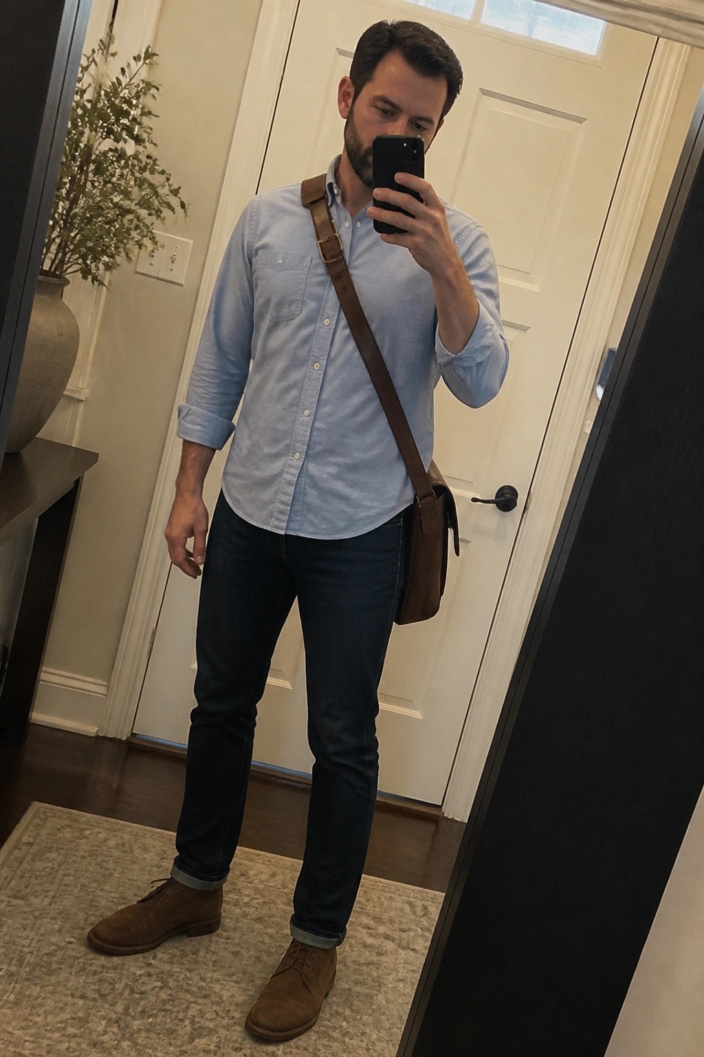 Bearded man in light blue shirt and dark jeans with brown messenger bag taking selfie