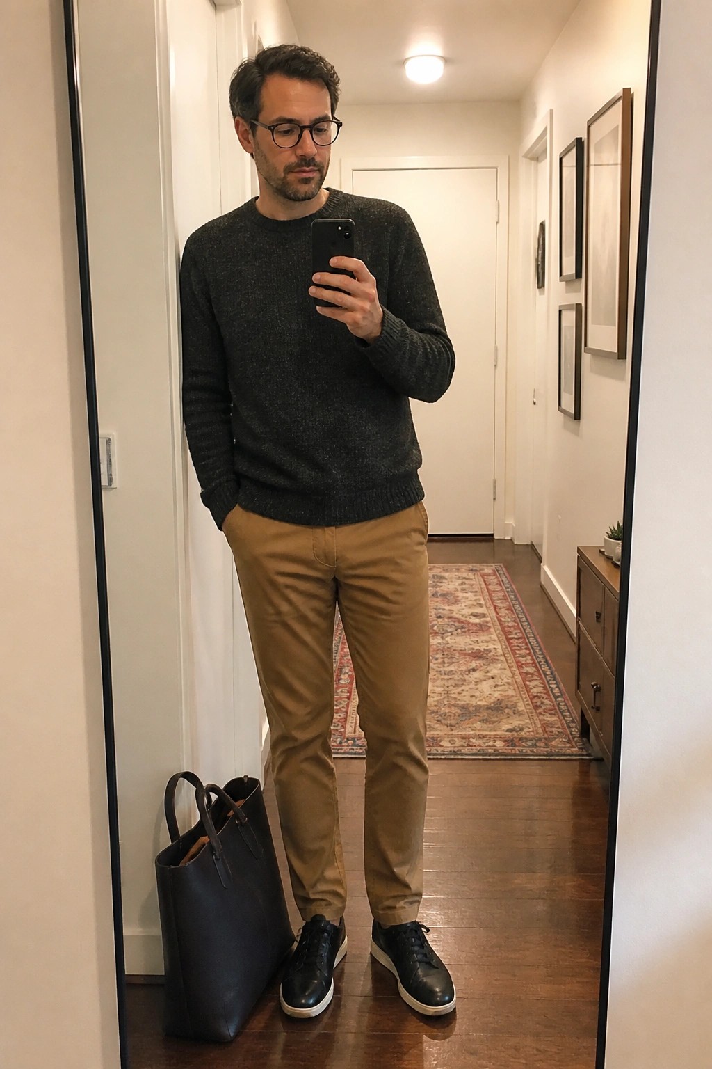 Man in dark sweater and tan pants taking mirror selfie beside black tote bag in hallway.