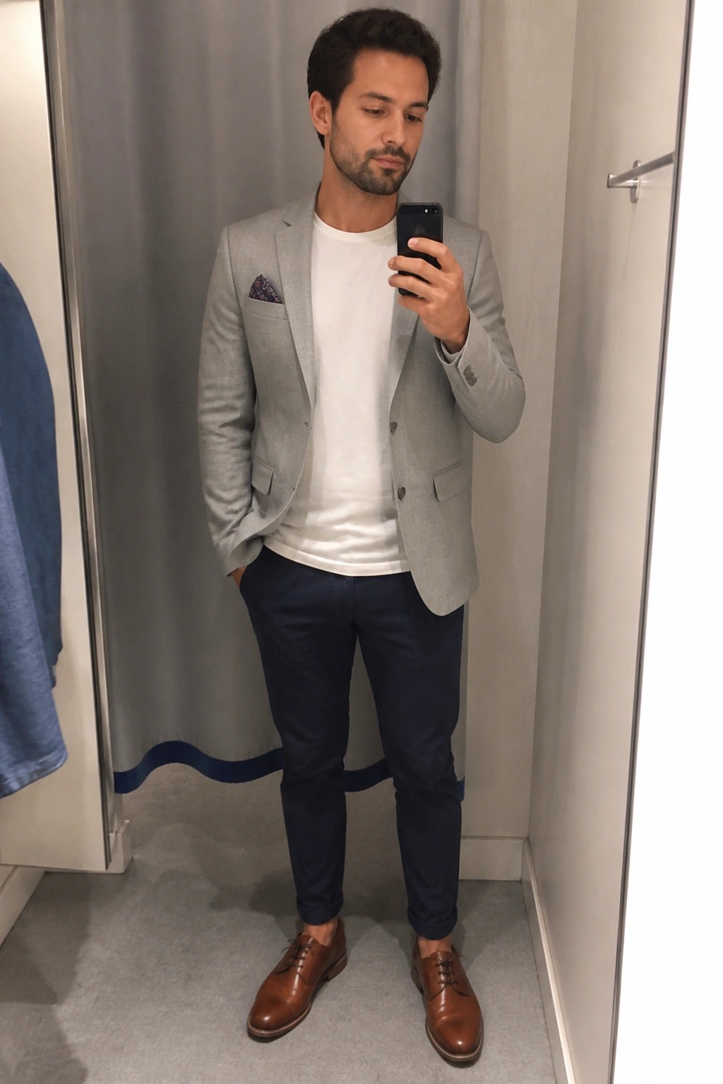 Man in gray blazer, white t-shirt, and navy pants taking mirror selfie in fitting room