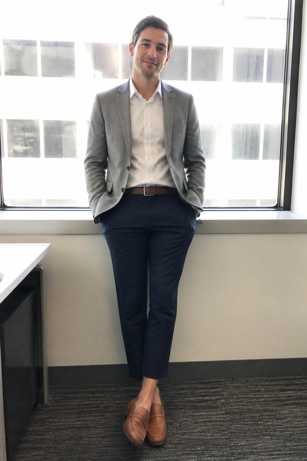 Man in gray blazer, white shirt, navy pants, and brown loafers by office window.