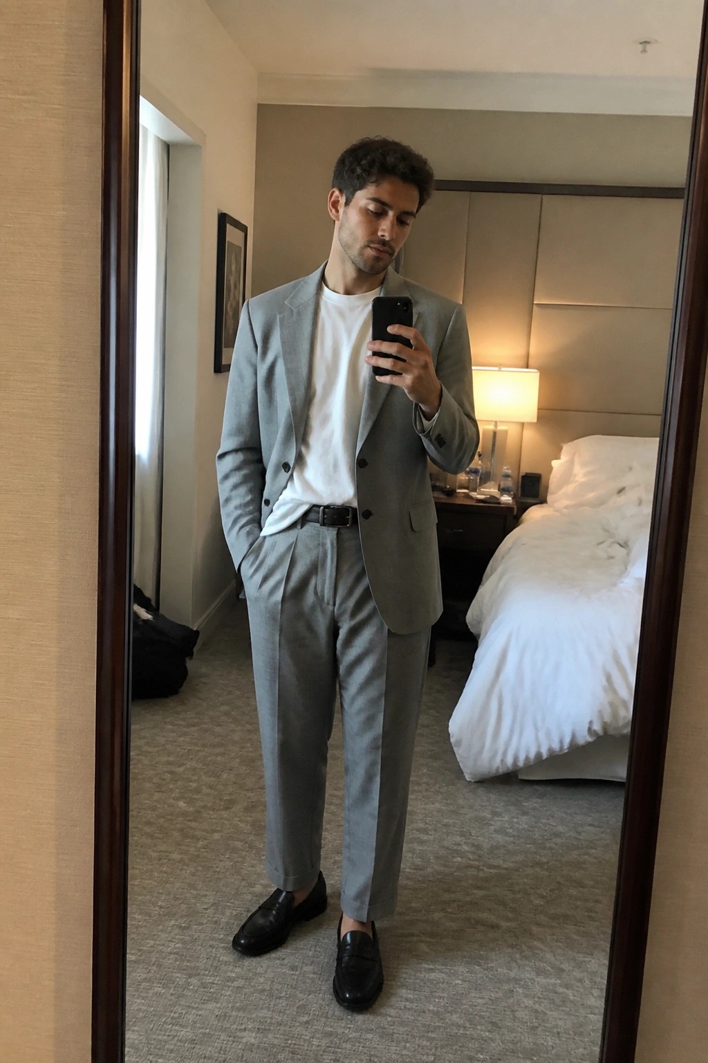 Man in light gray suit taking mirror selfie in hotel room with white bed