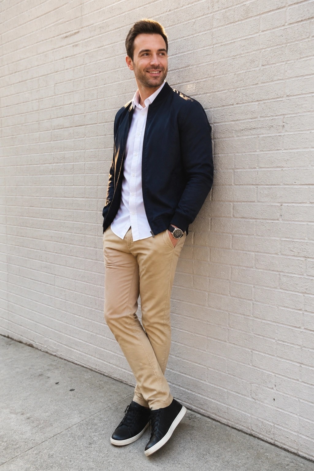 Smiling man in navy bomber jacket, white shirt, and khaki pants against brick wall.