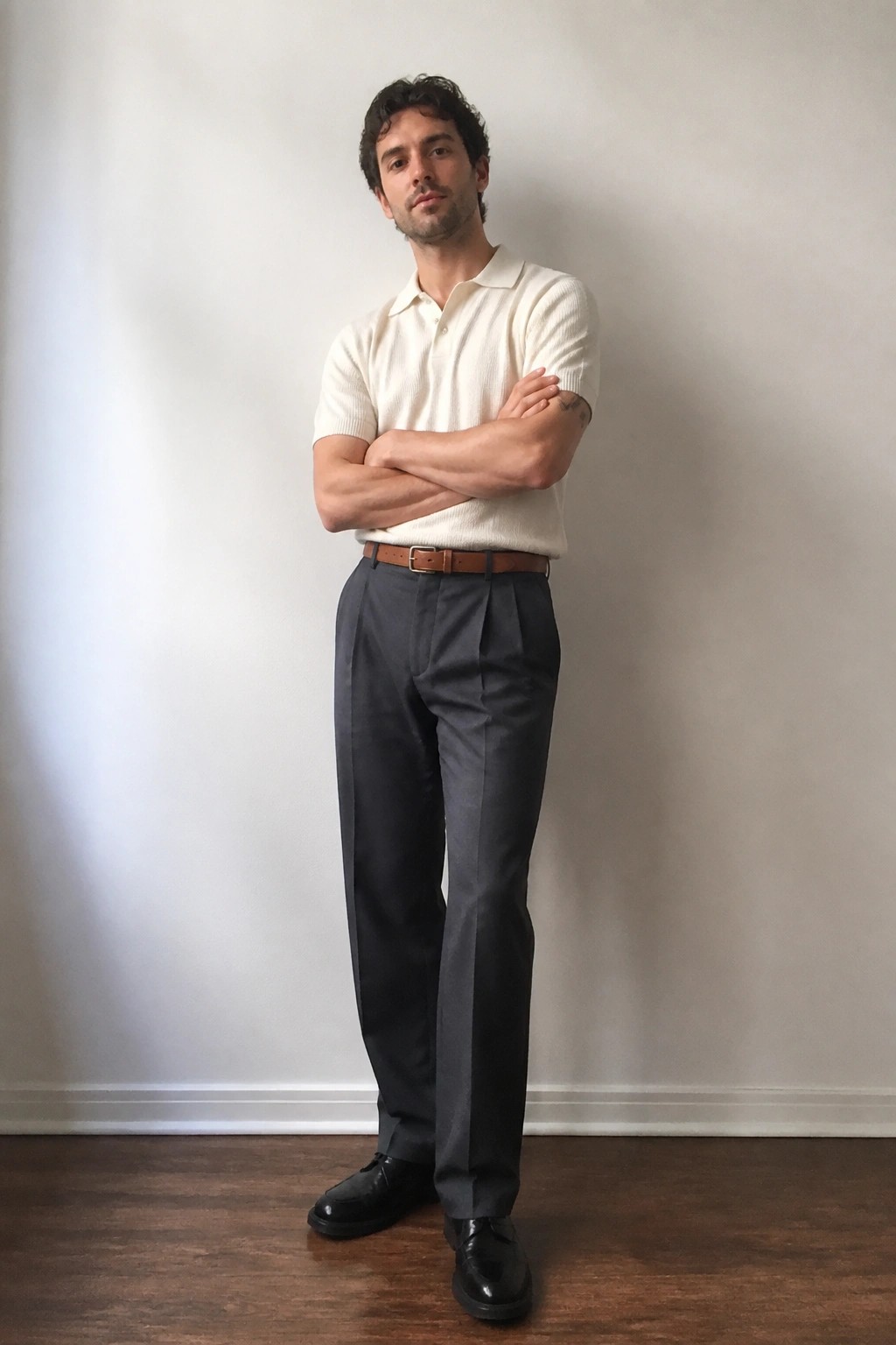 Man in cream polo shirt and gray pleated pants stands arms crossed