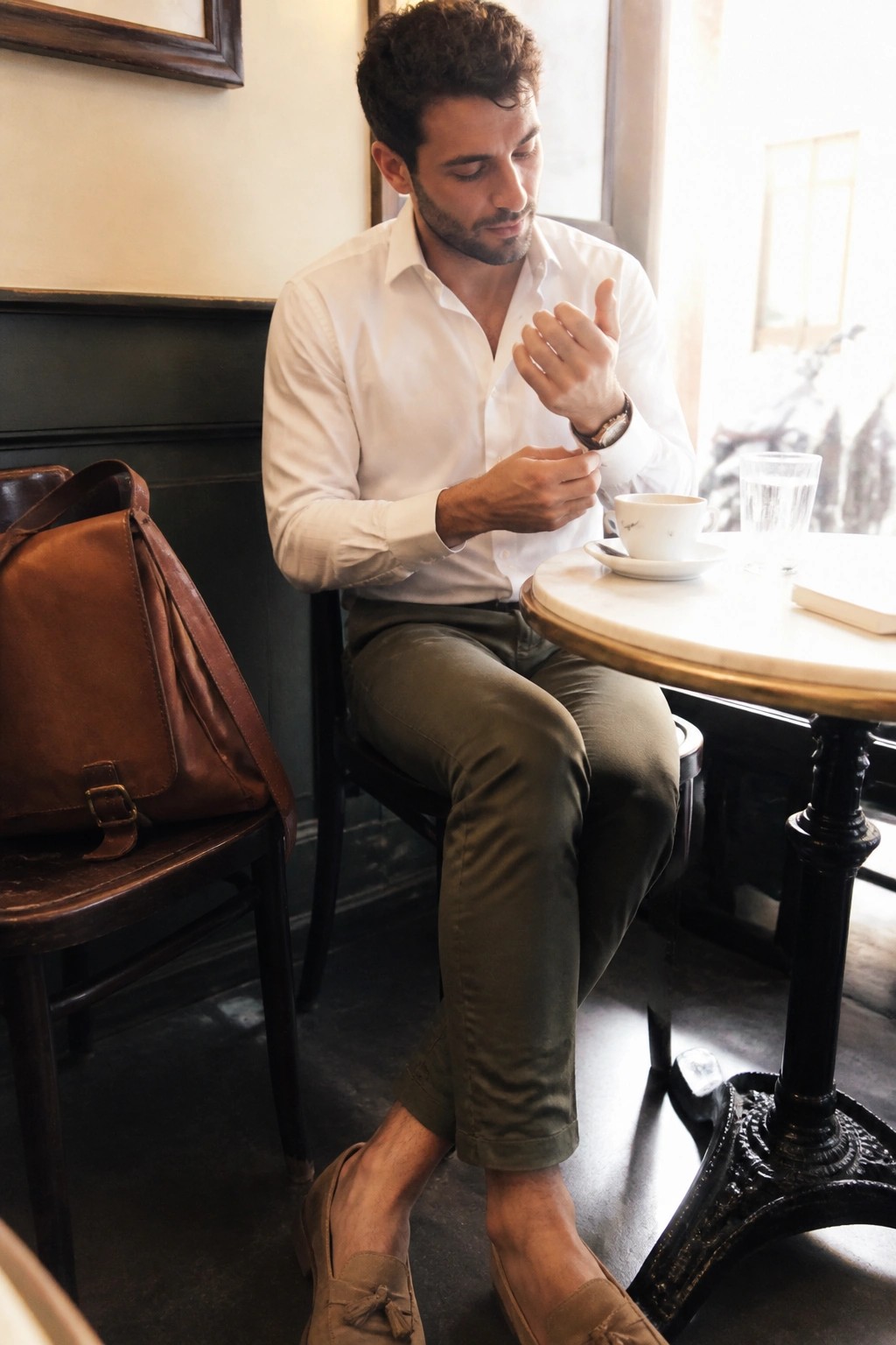 Man in white shirt and olive pants at marble cafe table with coffee and leather bag.