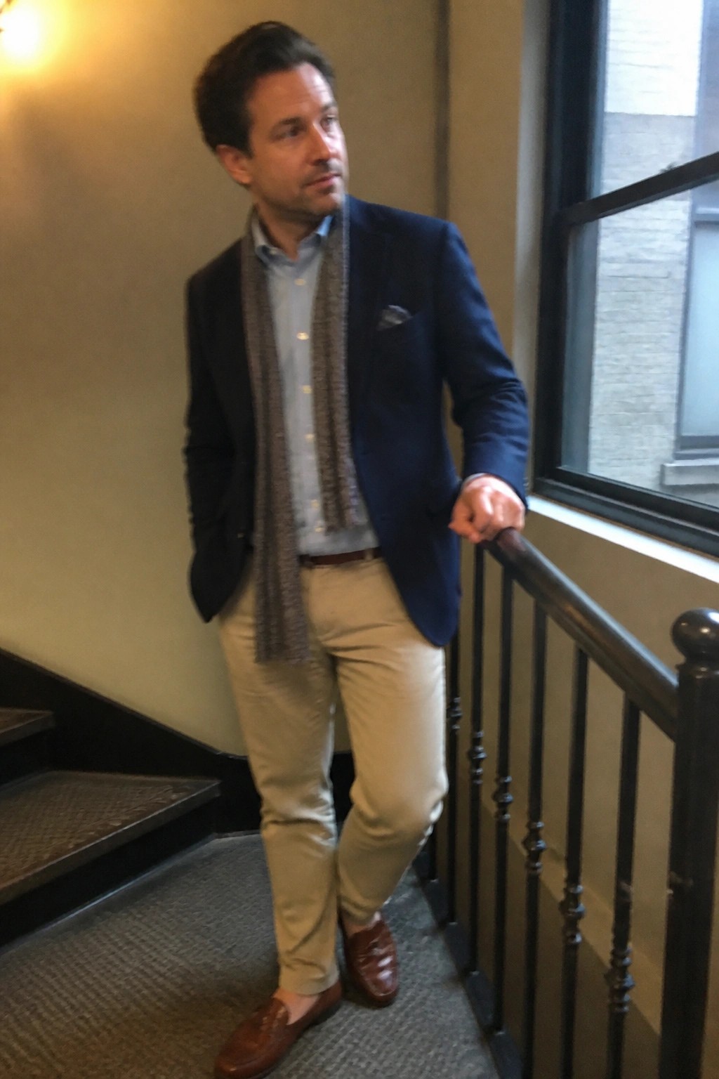 Man in navy blazer, gray scarf, and beige pants on indoor staircase