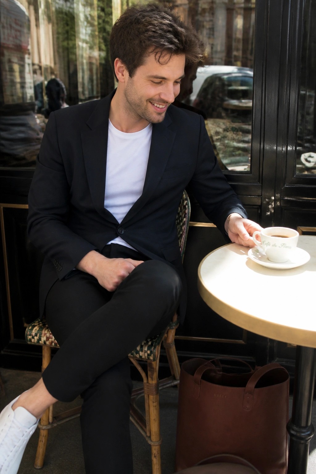Man wearing black blazer over white t shirt with black pants and white sneakers seated at cafe table with brown leather tote bag nearby.