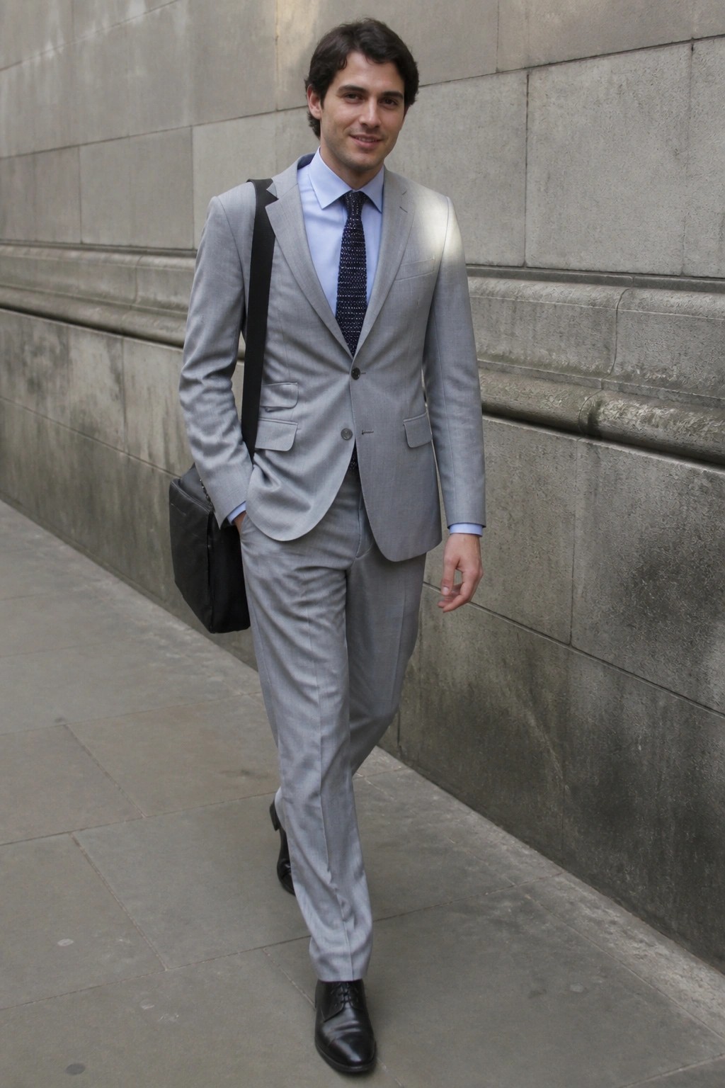 Man in light gray suit, blue shirt, patterned tie, with black shoulder bag.