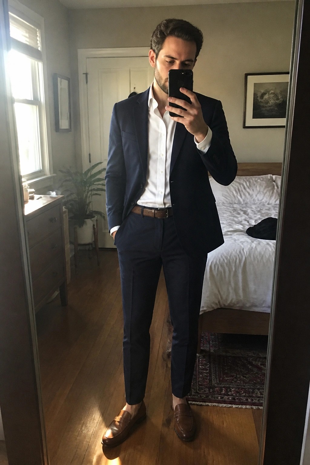Man in navy suit with white shirt and brown loafers taking bedroom mirror selfie