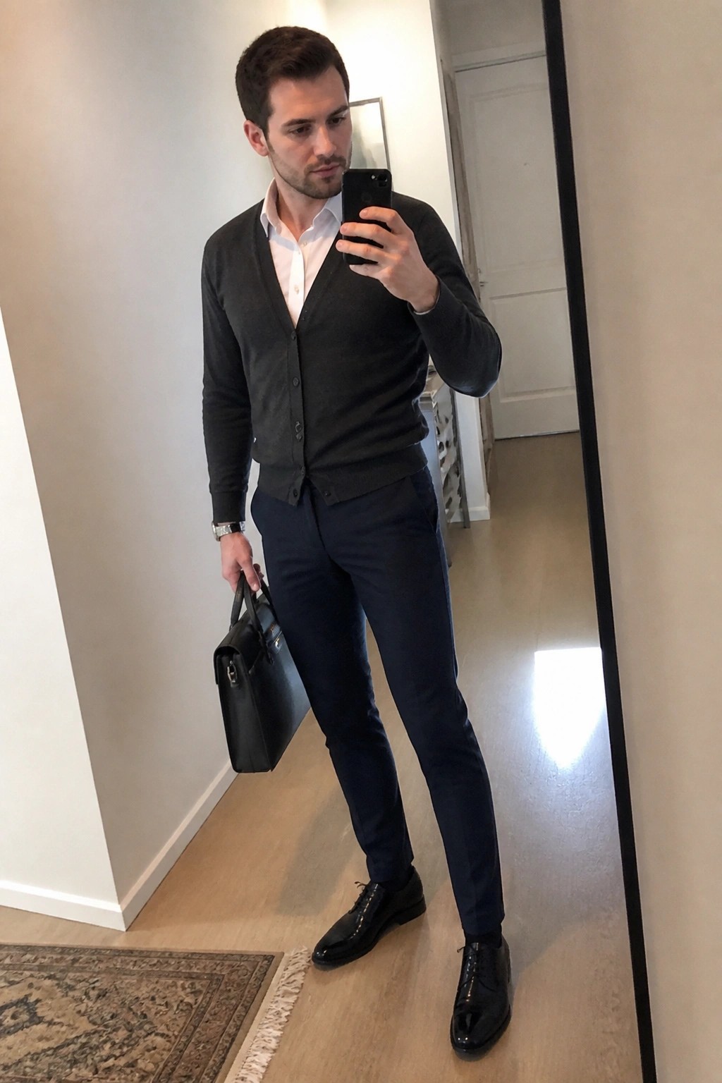 Man in gray cardigan and navy pants taking mirror selfie with black briefcase