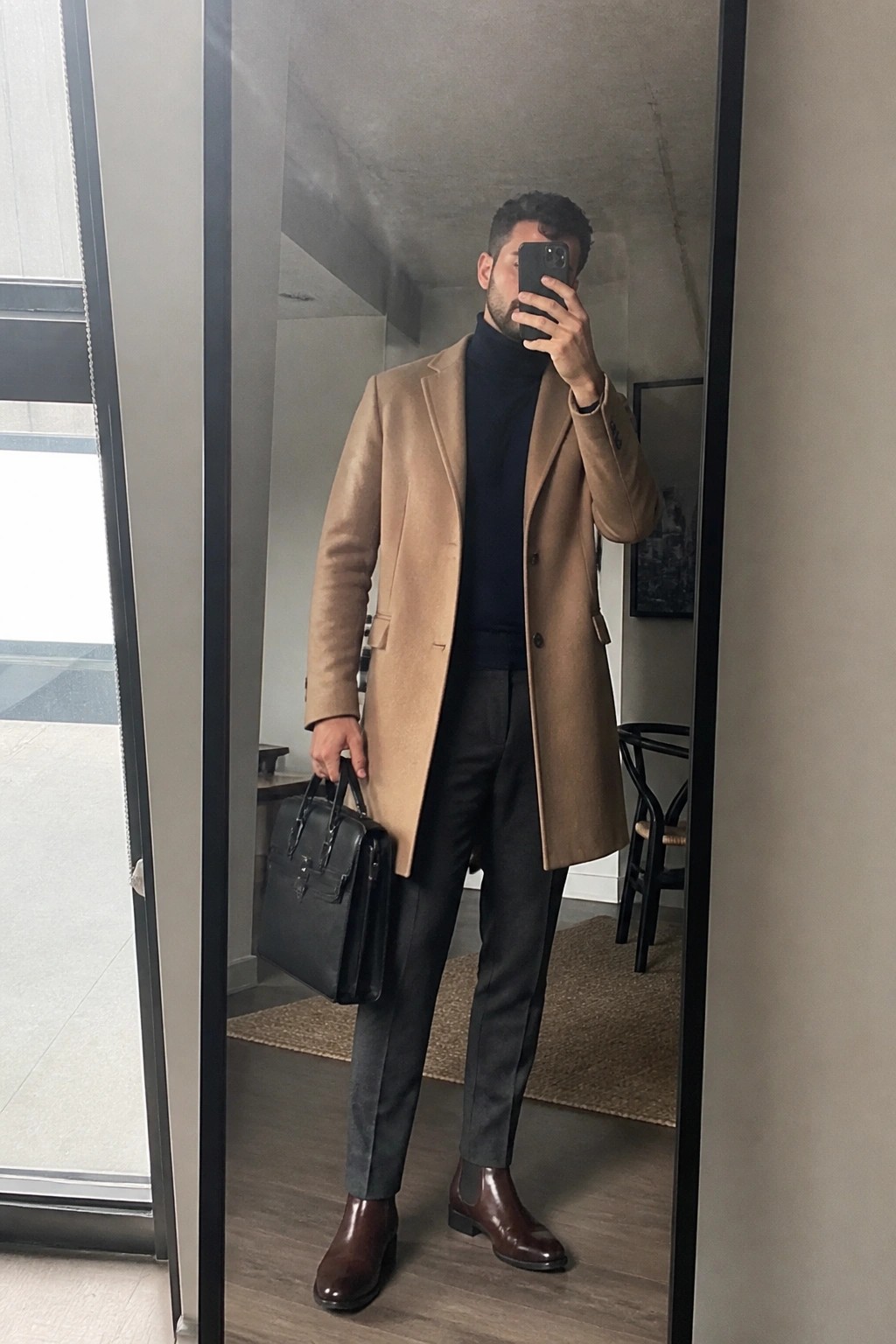 Man in tan overcoat and navy turtleneck holding black briefcase in mirror selfie