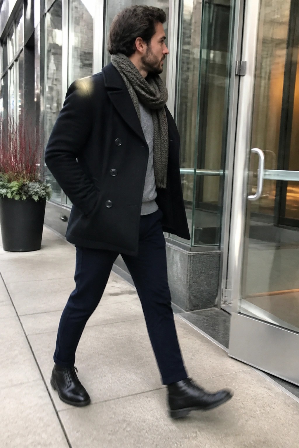 Man in black double breasted peacoat over gray sweater with gray knit scarf, navy pants and black boots standing outdoors.