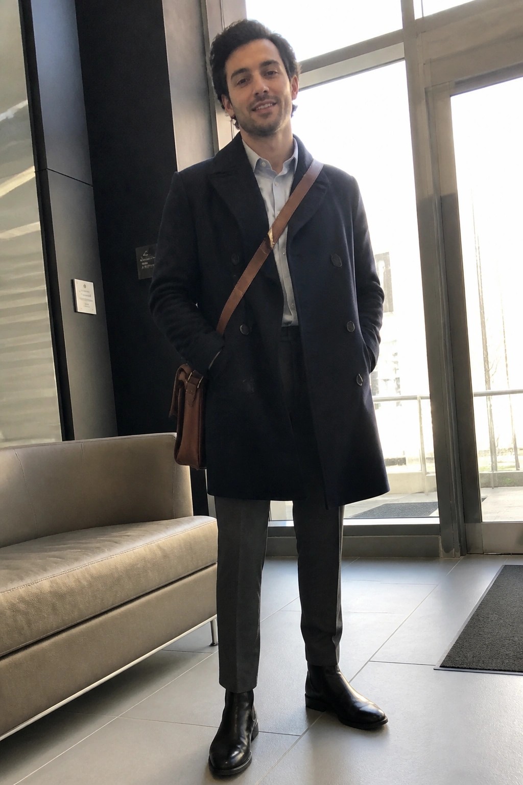Man in navy overcoat with brown leather bag stands in modern lobby