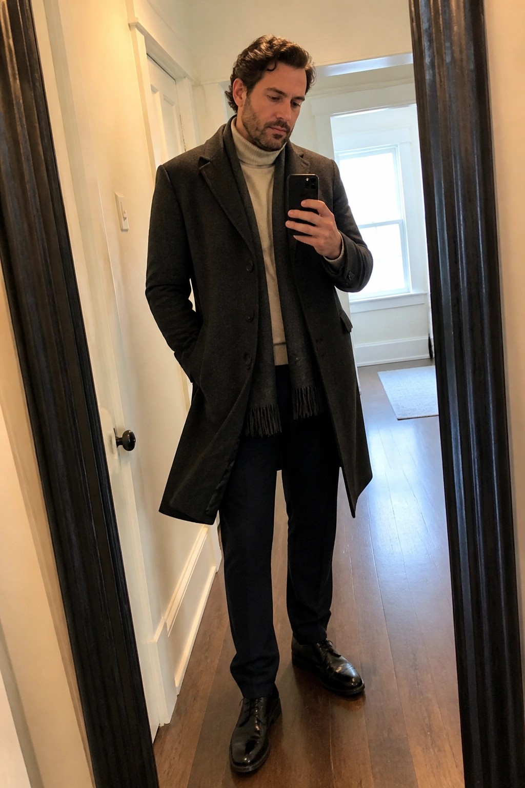Man in dark overcoat and beige turtleneck taking mirror selfie in hallway