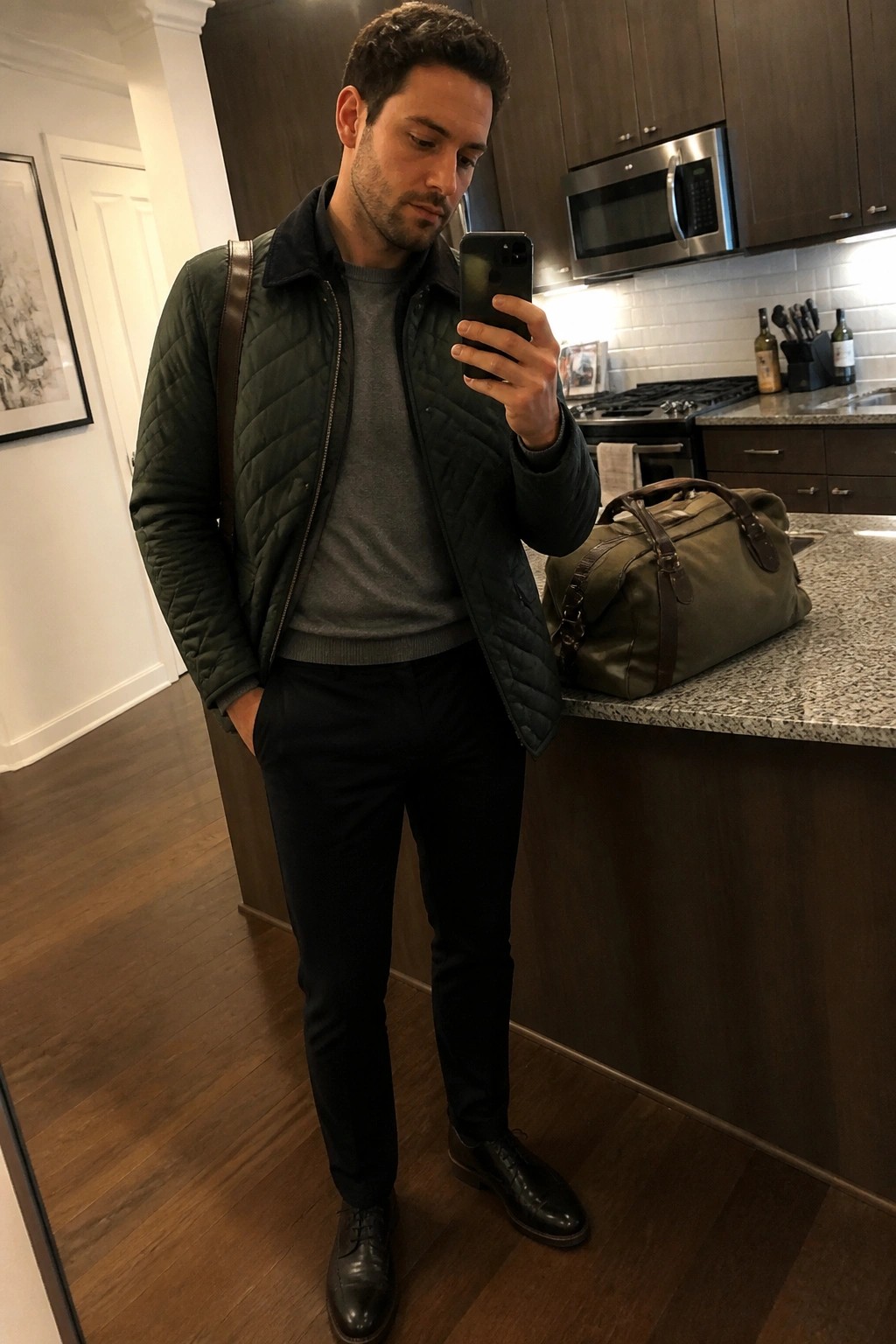 Man wearing dark green quilted jacket over gray sweater with black trousers and black dress shoes while holding a phone in a kitchen setting