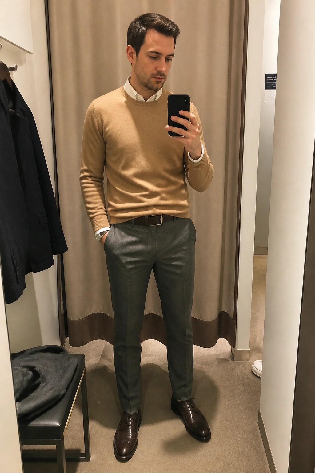 Man in tan sweater and gray pants taking mirror selfie in fitting room.