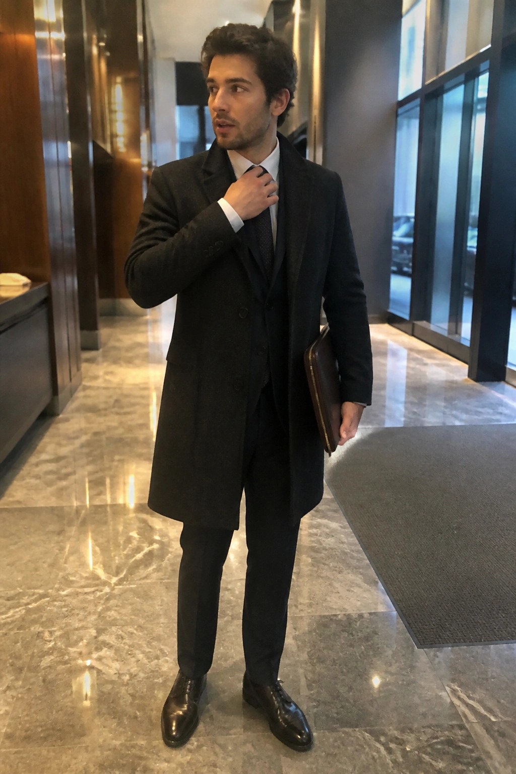 Man in dark overcoat and suit adjusting tie while holding leather portfolio in marble lobby