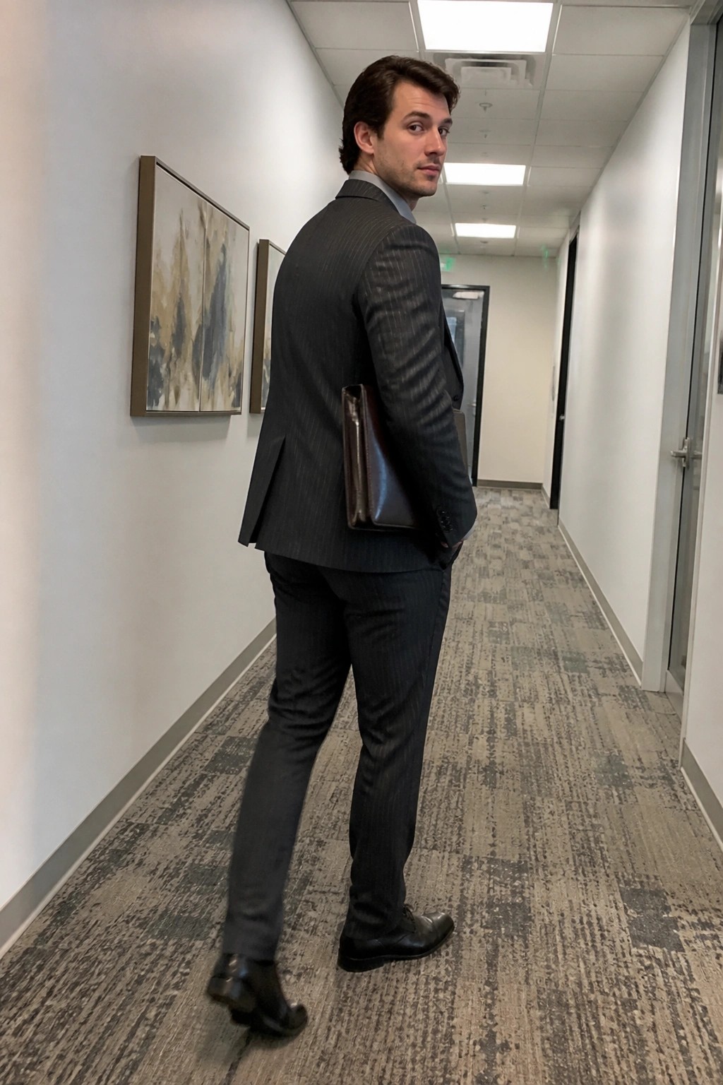 Man in dark pinstripe suit with leather portfolio looks back in office hallway