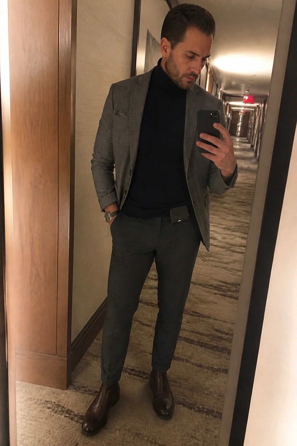 Man wearing a gray textured blazer over a navy turtleneck, paired with dark gray tailored trousers, brown leather Chelsea boots, and a silver watch, standing in a hallway while holding a phone.