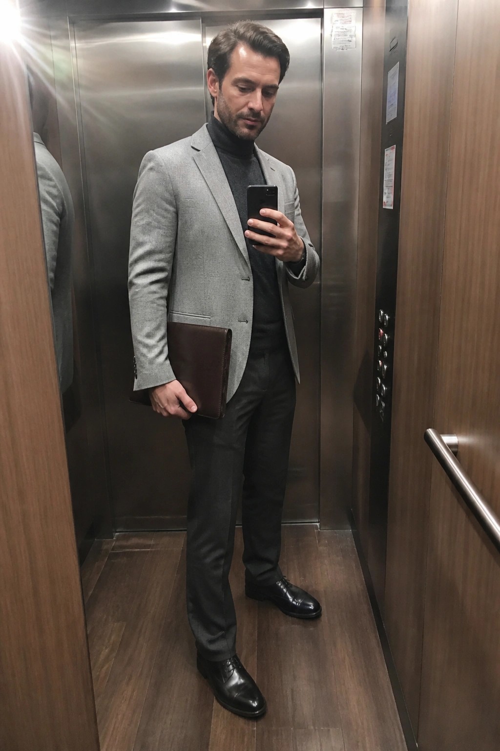 Man in gray blazer and turtleneck viewing phone while holding brown leather portfolio in elevator.