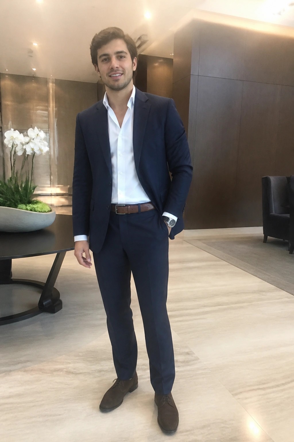 Man in navy suit with hand in pocket smiles in modern lobby.