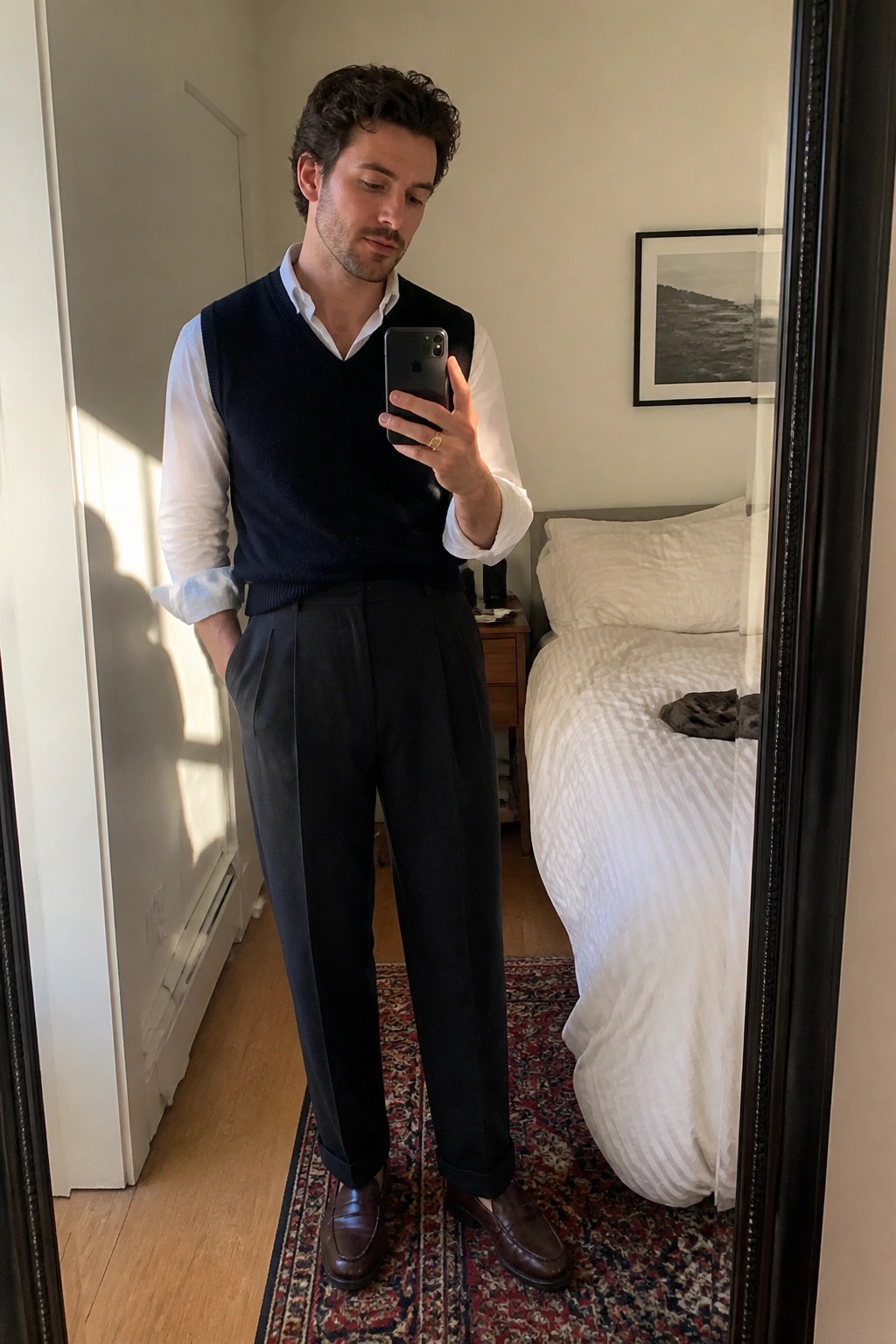 Man in navy vest over white shirt takes mirror selfie in bedroom