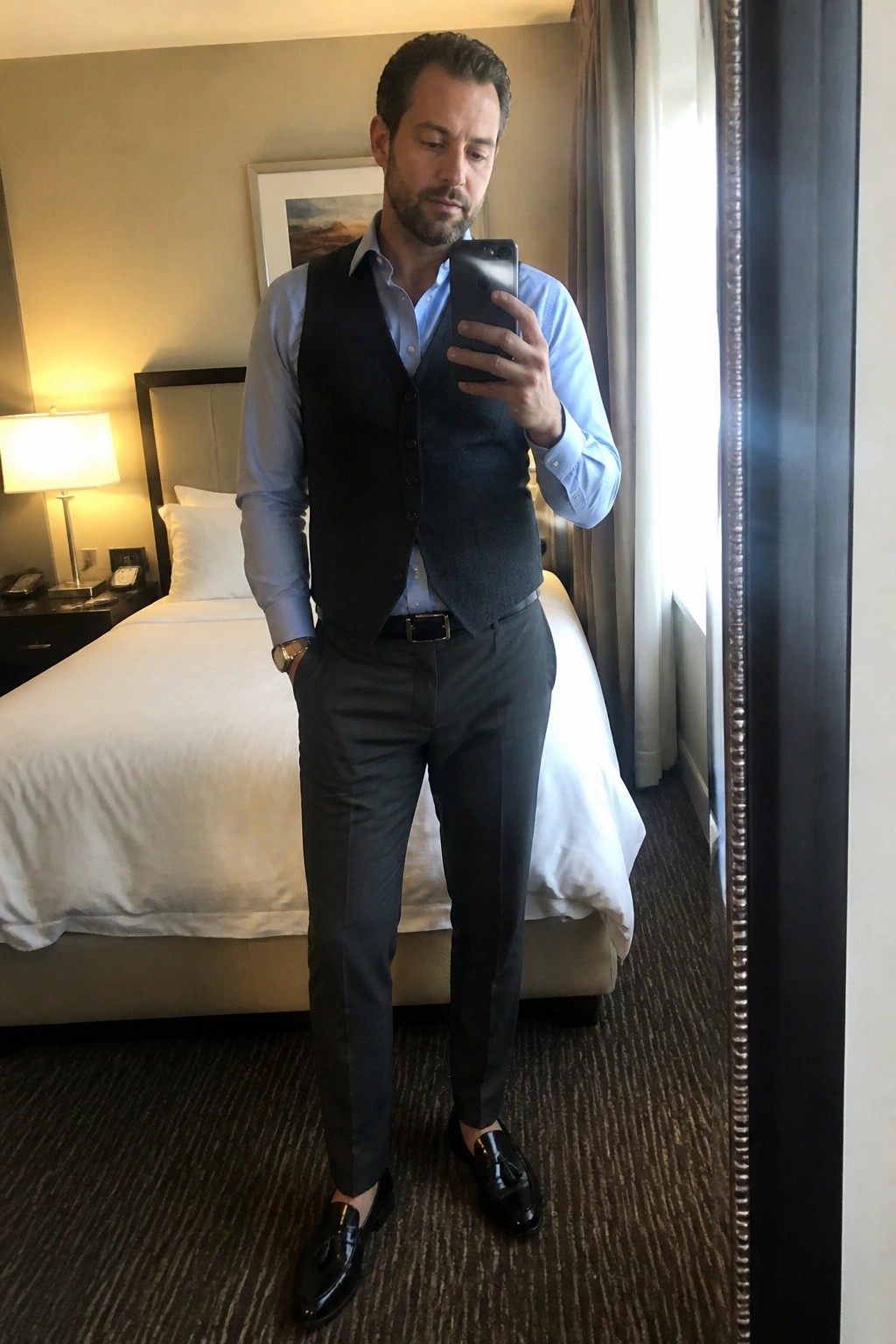 Man in blue shirt and vest taking mirror selfie in hotel room