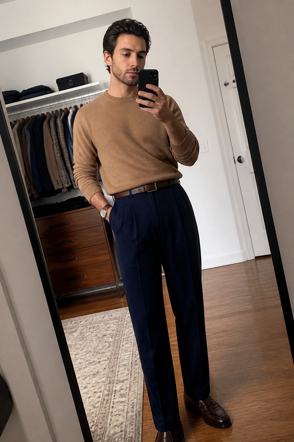 Man in tan sweater and navy pleated trousers taking mirror selfie with phone