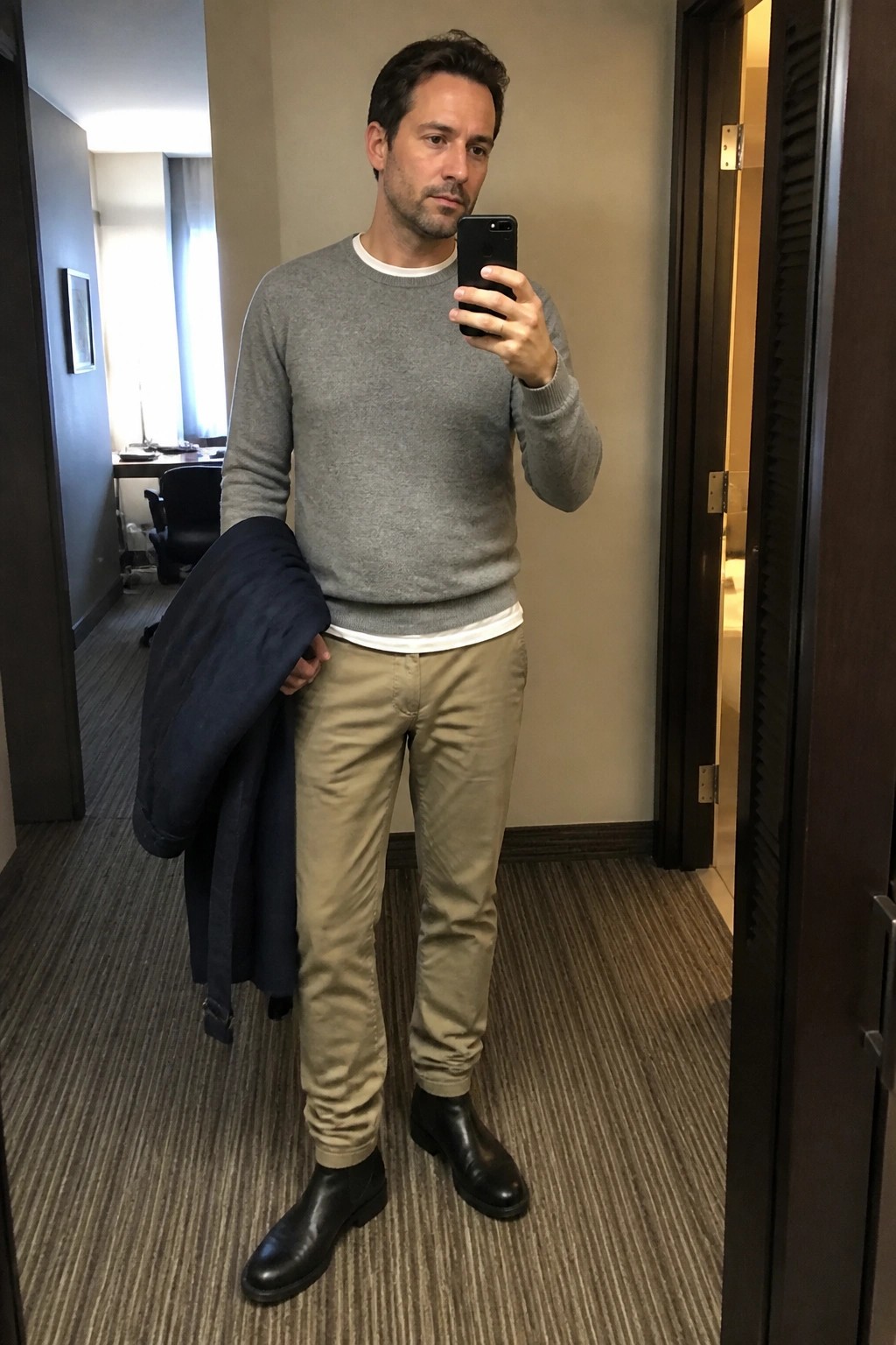Man in gray sweater and khaki pants takes mirror selfie while holding jacket
