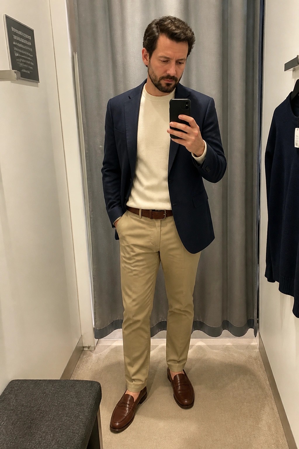 Man in navy blazer, cream sweater, and khaki pants taking mirror selfie with phone