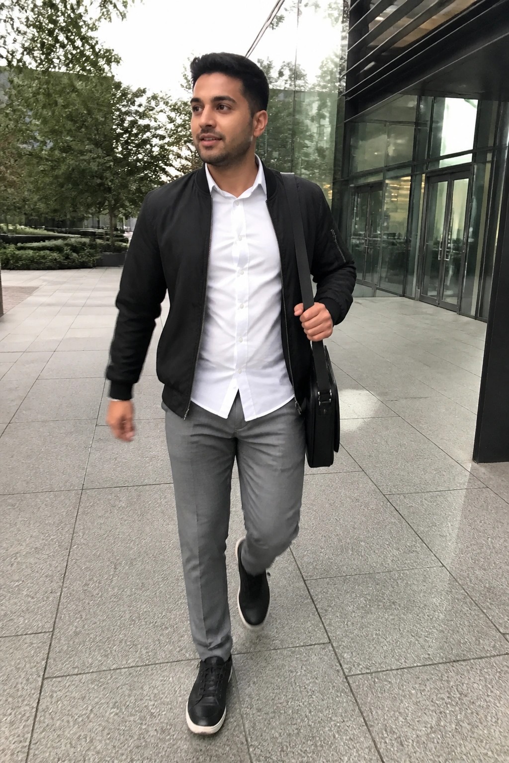 Man in black bomber jacket, white shirt, gray pants carrying black bag outdoors