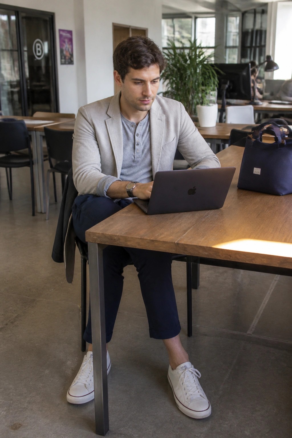 Man in beige blazer and white sneakers works on laptop at wooden table