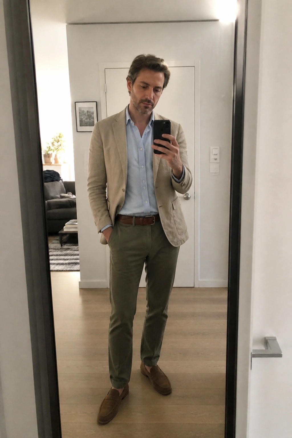 Man in beige blazer, blue shirt, and olive pants taking mirror selfie indoors