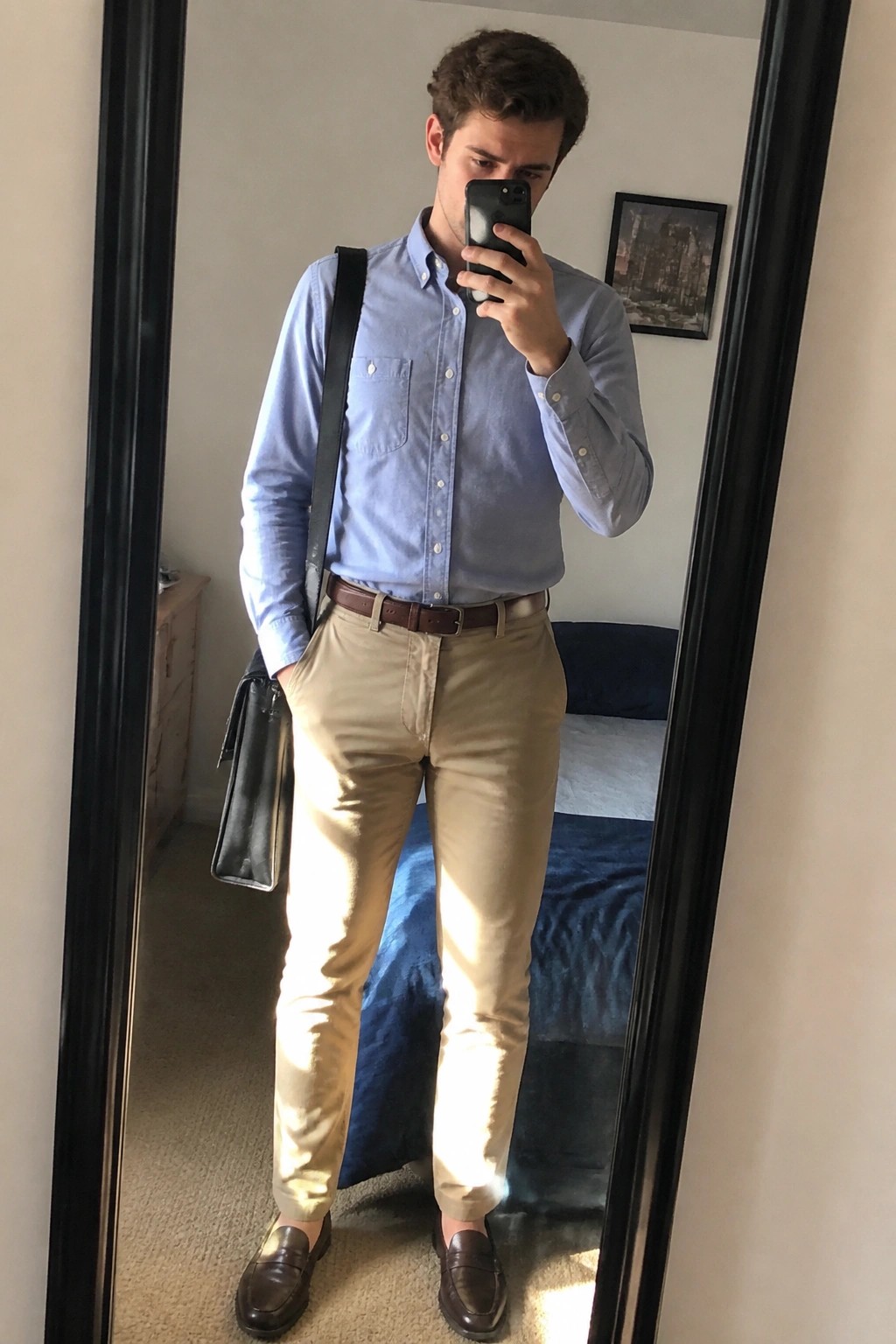 Man in light blue shirt and khaki pants taking mirror selfie with bag