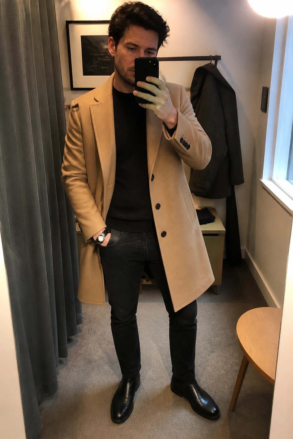 Man in tan overcoat over black sweater taking mirror selfie with phone
