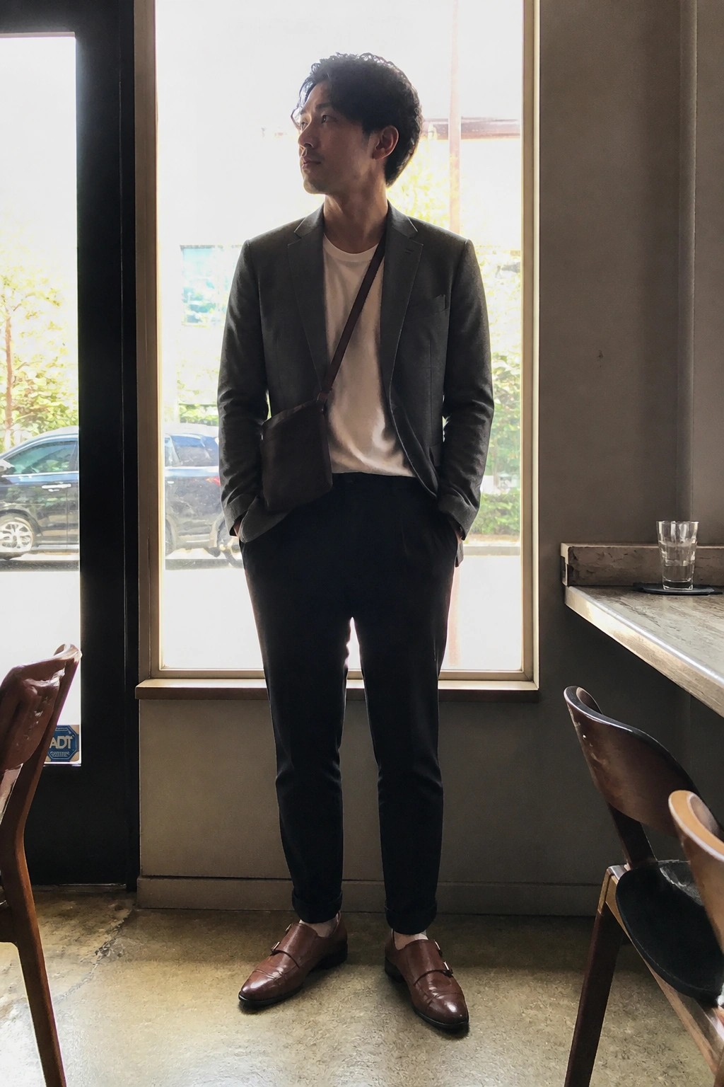 Man in gray blazer over white tee with brown bag stands by window.