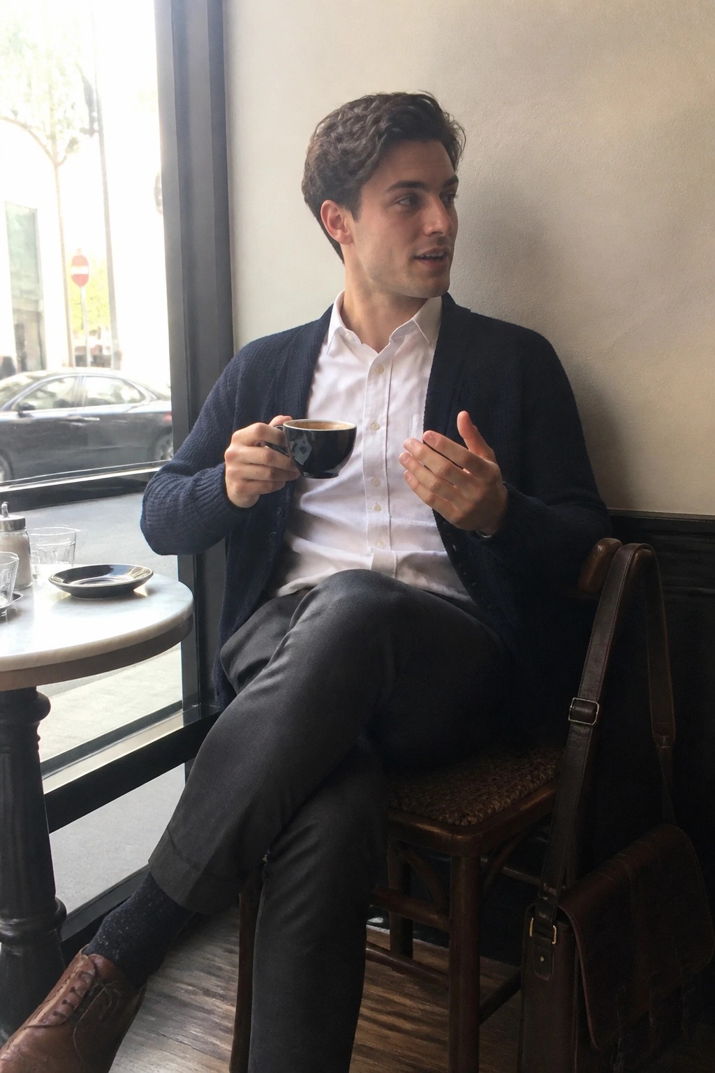 Young man in navy cardigan over white shirt holding coffee by cafe window
